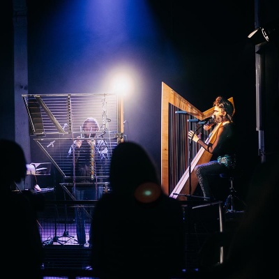 Hans Unstern und Simon Bauer an ihren 7 selbstgebauten experimentellen Harfen. Perkussionelemente im Hintergrund hängen an Seilen von der Decke. Stimmungsvolles Konzertlicht in blau. 