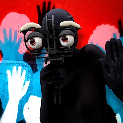 Eine schwarze Figur hält sich eine mechanische Maske vors Gesicht und steht gleichgültig winkend vor einer Projektion. Die Hintergrundprojektion zeigt eine Menschenmenge in den Farben weiß, schwarz, türkis und rot. Die Menschen strecken ihre Arme jubelnd 